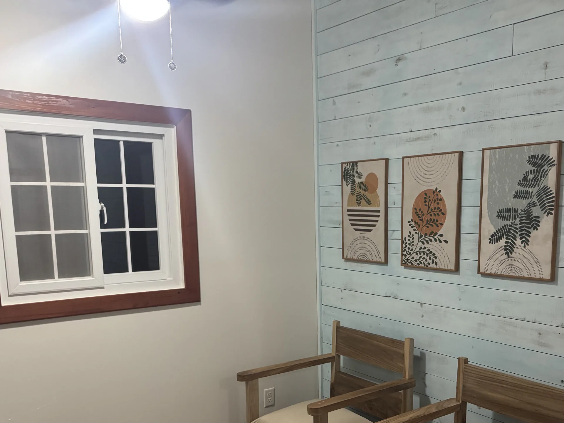 Sitting room with two teak armchairs, three prints on a whitewashed shiplap wall, and a window